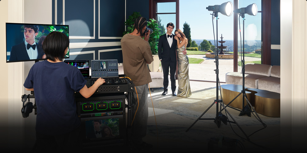 Behind-the-scenes setup: a couple in formal wear pose by a large window overlooking the sea, while crew operate video gear and lighting nearby.
